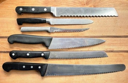 Six different styles of serrated knife lined up on a wooden cutting board.