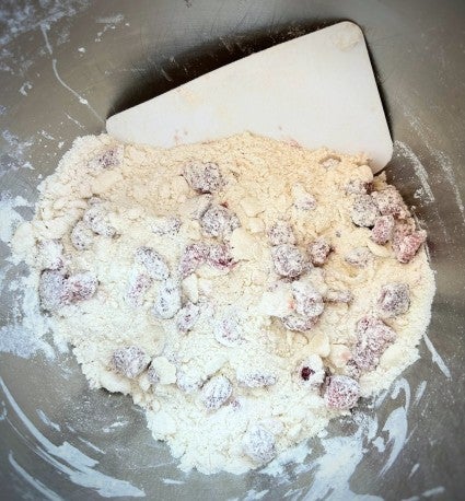 Diced fresh strawberries mixed with flour and other dry ingredients in a stainless steel bowl, with a dough scraper. 